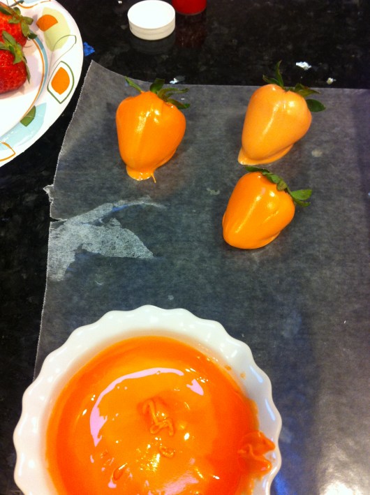 Melt the candy coating in a ramekin deep enough to submerge your berries; then drop enough of the orange candy dye into the melted coating to turn it a "carrot orange" color; dip strawberries until fully coated other than the leaves and place finished onto some wax paper; finally, push leaves back into place atop the berries to "hide" any exposed red spots (the wet chocolate also help hold the leaves down in place).